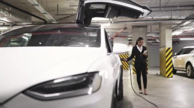 Halka açık garajda saf elektrikli aracı doldururken Afrikalı Amerikalı bir kadının telefonda konuşmasının düşük açılı görüntüsü. Ofis dışındaki şirket meseleleriyle ilgili kararlar veren resmi kıyafetli meşgul bir işçi..