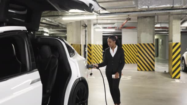 アフリカ系アメリカ人女性は 公共のガレージで純粋な電気自動車を満たしながら電話で話します オフィスの外で会社の問題を決定する正式な衣装の忙しい労働者 — ストック動画