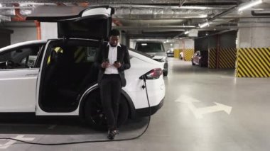 Afro-Amerikan bir adamın EV dolum sırasında içki içtiği ve telefon görüşmelerini kontrol ettiği bodrum katındaki otoparkı geniş açıdan görebiliyoruz. İş yerinde elektrikli otomobili çalıştırmak için kahve molası veren rahat bir yetişkin..