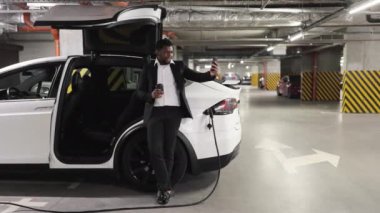 Takım elbiseli çekici bir adamın e-arabanın yanında kahve içerken akıllı telefondan fotoğraf çekmesi. Afrika kökenli Amerikalı çalışanlar park yerinde dinlenme anı yakalıyorlar..