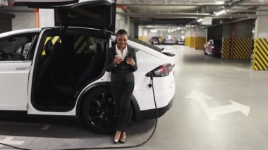 Kahveli Afro-Amerikan işçinin cep telefonuyla EV 'nin yanında konuştuğu alt kattaki otoparkı geniş açıdan görebiliyoruz. Kendine güveni tam bir bayan mola verirken şarj istasyonunu pilli araba için kullanıyor..