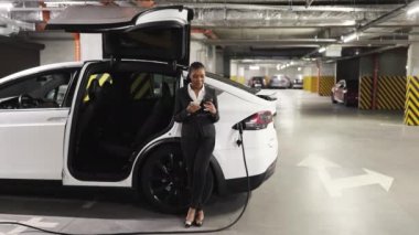 Kahveli Afro-Amerikan işçinin cep telefonuyla EV 'nin yanında konuştuğu alt kattaki otoparkı geniş açıdan görebiliyoruz. Kendine güveni tam bir bayan mola verirken şarj istasyonunu pilli araba için kullanıyor..