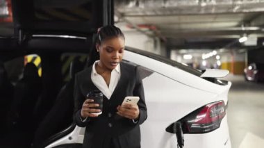 Afrika kökenli Amerikalı kadının e-araba arka planında hazır içkiyle telefon kamerasına gülümsemesini yakından izleyin. EV 'nin yakınındaki yeraltı otoparkında kendi portresini yapan neşeli bir çalışan..