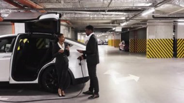 E-otomobili şarj noktasına bağlarken iki resmi kıyafetli kişinin tartışmaya karıştığı tam boy görüntüsü. Aletleri olan Afrikalı takım arkadaşları ve bodrum katındaki garajda kahve paylaşımı önerileri..