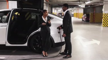 EV portundaki elektrik bağlantısını takan erkek meslektaşına mobil ekran görüntüsü gösteren yemek kupalı heyecanlı bir bayan. Afro-Amerikan iş adamları garajdaki şirket sorunları hakkında bilgi paylaşıyorlar..
