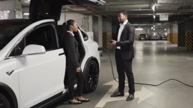 Afrika kökenli Amerikalı kadın ve erkek beyaz bir e-arabanın yanında el sıkışıyorlar. Şarj tabancası limana bağlı. Kalifiye ticari ortaklar yeraltı otoparkında anlaşmaya varılan iş anlaşmasını mühürlüyor.