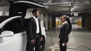 İş elbiseli çekici Afrikalı kadın yeni elektrikli otomobilin anahtarlarını yer altı otoparkındaki erkek alıcıya veriyor. Profesyonel satıcı EV ücret noktasında araba satın alma sürecini tamamlıyor.