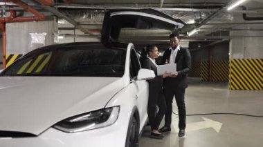 Portatif bilgisayarlı Afrikalı erkek ve kadın garajdaki elektrikli otomobil kapısının yanında duruyor. Araştırma sonuçlarıyla ilgili kurumsal diyaloğu yürüten etkili iş arkadaşları kapalı alanlarda.