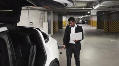 Resmi takım elbiseli gülümseyen orta yaşlı bir adam elinde bilgisayarla EV şarj kablosu takıyor. Afrikalı Amerikalı girişimci ofisten ayrıldıktan sonra elektronik iletişimi sürdürüyor.