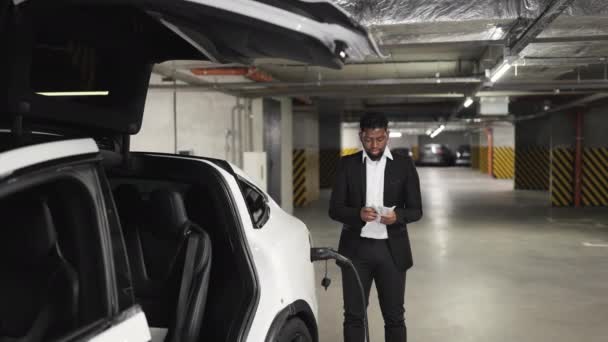 ビジネスススーツのアフリカの男性大人は 屋内充電ポイントでバッテリー駆動車両を供給しながら 現金で親指をスーツ Ev充電のための最近の支出を思い出させるハンサムな駐車顧客 — ストック動画