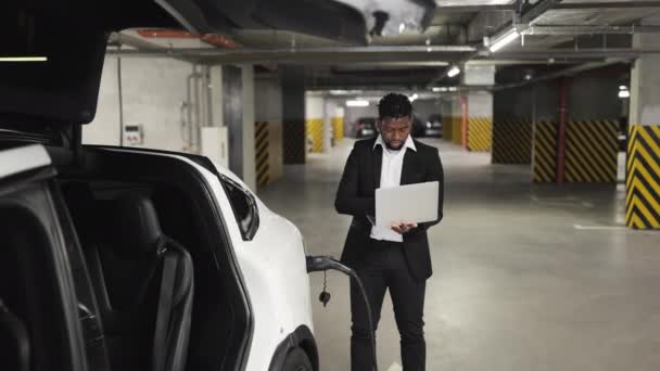 Evの公共駐車場でポータブルラップトップを活用する正式な衣装でアフリカのビジネスマンの選択的な焦点 Car近くのオフィス壁を越えてプロの活動を行う資格のある従業員 — ストック動画