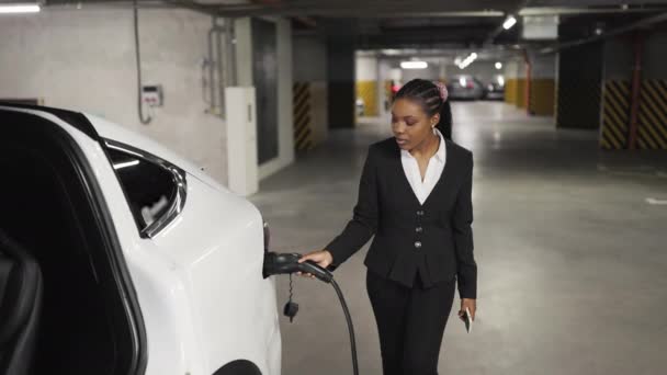 リラックスしたアフリカ系アメリカ人女性の肖像画は 駐車場に充電器銃を入れています スマートフォンでプロの起業家がビジネス旅行のための自動準備を取得 — ストック動画