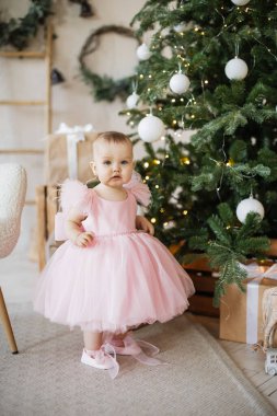 Beyaz, sevimli bebek Noel ağacında süslenmiş çelenge dokunuyor ve parmakları içeride. Stüdyonun köşesinde yeni yıl havasında şık güzel bir kız duruyor..