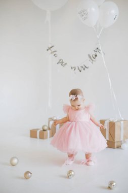 Stüdyoda hediyeler ve balonların arasında dikilirken şık bir tatil elbisesiyle ilk doğum gününü kutlayan sevimli küçük kız. Çocukluk ve kutlama kavramı.