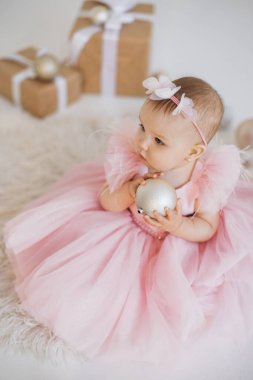 Festivale uygun giyinmiş küçük güzel prenses süslemeli stüdyoda elinde beyaz topla oturuyor. Güzel saç bandı takmış çekici bir bebek. Bakıyor ve doğum günü partisinin tadını çıkarıyor..