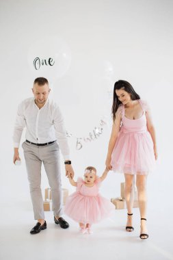 Sevimli pembe elbiseli sevimli küçük kızlarıyla el ele tutuşan mutlu genç ebeveynler. Beyaz erkek ve kadın sevgili çocuğun ilk doğum gününü stüdyoda dekor ve hediyelerle kutluyorlar..