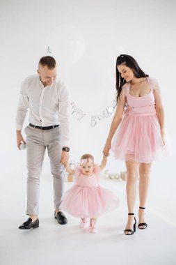 Sevimli pembe elbiseli sevimli küçük kızlarıyla el ele tutuşan mutlu genç ebeveynler. Beyaz erkek ve kadın sevgili çocuğun ilk doğum gününü stüdyoda dekor ve hediyelerle kutluyorlar..