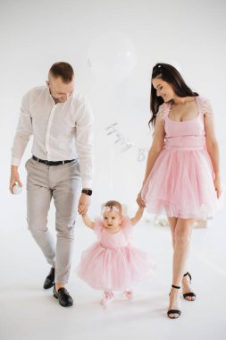 Sevimli pembe elbiseli sevimli küçük kızlarıyla el ele tutuşan mutlu genç ebeveynler. Beyaz erkek ve kadın sevgili çocuğun ilk doğum gününü stüdyoda dekor ve hediyelerle kutluyorlar..