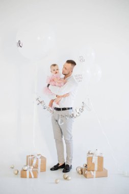 Doğum günü süslemeleriyle stüdyoda dikilirken neşeli pembe elbiseli şirin kızı kucaklayan mutlu genç babanın tam boy portresi. Ebeveynlik ve kutlama konsepti.