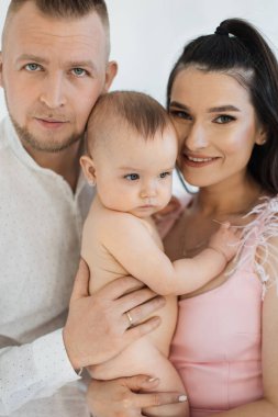 Güzel bir eşin kucağında, kollarında kıyafetleri olmadan güzel bir bebek tutması. Mutlu aile kameraya bakıyor ve güzel ışıklandırılmış stüdyoda harika vakit geçiriyorlar..