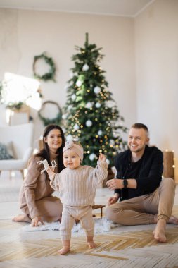 Güzel bir Noel ağacının yanında oturan beyaz eşiyle ilgilenmek ve şık örgü örülmüş şirin küçük kızının ilk adımlarının tadını çıkarmak. Kış tatilinde mutlu aile anları.