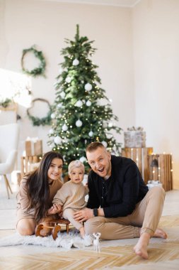 Neşeli, genç ebeveynler, parlak Noel ağacının yanında oturup oyuncak kızakla sevimli bir kızı kucaklıyorlar. Üç kişilik beyaz bir aile rahat evlerinde şenlik düzenliyor..