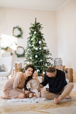 Neşeli, genç ebeveynler, parlak Noel ağacının yanında oturup oyuncak kızakla sevimli bir kızı kucaklıyorlar. Üç kişilik beyaz bir aile rahat evlerinde şenlik düzenliyor..