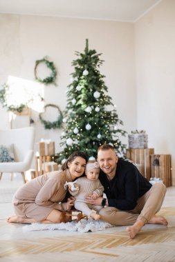 Neşeli, genç ebeveynler, parlak Noel ağacının yanında oturup oyuncak kızakla sevimli bir kızı kucaklıyorlar. Üç kişilik beyaz bir aile rahat evlerinde şenlik düzenliyor..