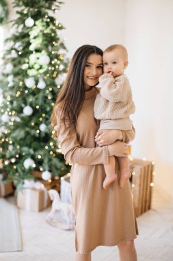 Çekici beyaz bir kadının sevimli bir kızı ellerinin arasında tutması ön planda. Güzel anne şık bej elbiseler giyip küçük kızla eğleniyor. Noel ağacının arka planı.