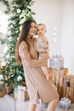 Çekici beyaz bir kadının sevimli bir kızı ellerinin arasında tutması ön planda. Güzel anne şık bej elbiseler giyip küçük kızla eğleniyor. Noel ağacının arka planı.