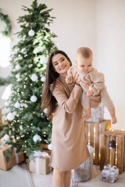 Çekici beyaz bir kadının sevimli bir kızı ellerinin arasında tutması ön planda. Güzel anne şık bej elbiseler giyip küçük kızla eğleniyor. Noel ağacının arka planı.