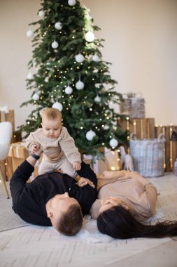 Neşeli beyaz ebeveynler yerde küçük bir bebekle yatıyorlar ve evde Noel atmosferinin tadını çıkarıyorlar. Birlikte oynayan ve neşeli duygular ifade eden üç kişilik güzel bir aile..