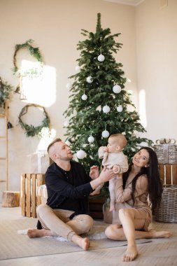 Şık giyinen neşeli beyaz eş güzel Noel ağacının yanında oturup sevimli kızlarına tezahürat yapıyor. Genç ebeveynler, kış tatillerinde çocuklar için neşeli bir ortam yaratıyorlar..