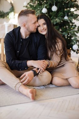 Yakışıklı bir gencin, sıcak oturma odasında birlikte otururken sevgili karısını öpüp kucaklamasının portresi. Arka planda parlayan Noel ağacı. Bayram kutlamalarında aile anları.