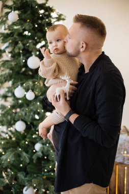 Güzel beyaz bir baba, küçük sevimli kızının yanağını öpüyor. Arkaplanda parlak çelenklerle Noel ağacı var. Sıcak örgü süveter giymiş, tatlı bir kız..