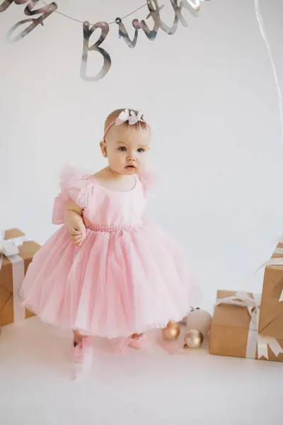お祝いの装飾で飾られたスタジオで一人で立っている最高のパーティードレスを着て遊び心のある少女 誕生日パーティーを祝う誠実な感情を持つ慎重な赤ちゃんの少女を魅了する — ストック写真