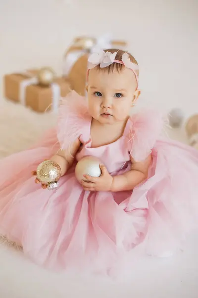 Small beautiful princess dressed in festive wear sitting with white ball in hands in decorated studio. Attractive pretty toddler with cute headband looking aside and enjoying birthday party.