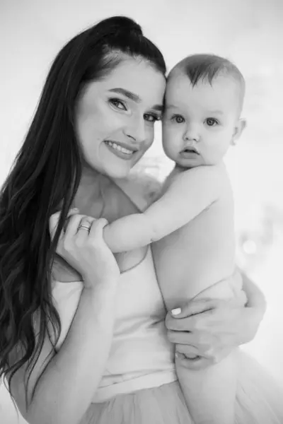 Black and white portrait of mom and nude beautiful daughter expressing pure and unconditional love between them. Charming woman standing at studio with little adorable toddler on hands.