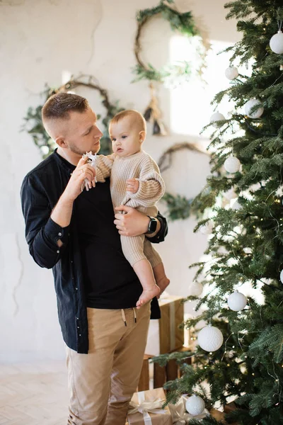 Portrait of happy young dad smiling and looking at camera while holding pretty little infant on hands near shiny Christmas tree. Caucasian family at home during winter holidays.