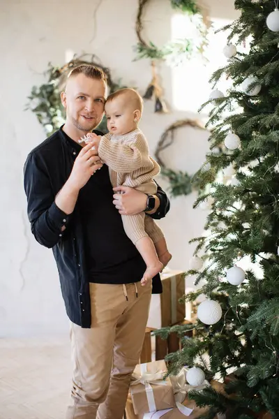 Portrait of happy young dad smiling and looking at camera while holding pretty little infant on hands near shiny Christmas tree. Caucasian family at home during winter holidays.