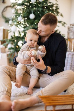 Yakışıklı genç bir baba kucağında küçük kızını tutarken ve Noel ağacı süslemelerini gösterirken yerde oturuyor. Önemseyen baba, küçük sevimli çocuğa baktığında mutluluğu hisseder..