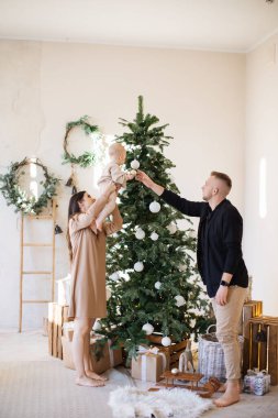 Dost canlısı beyaz aile Noel ağacı ve yeni yıl köşesini birlikte süslüyor. Şık giyinmiş pozitif ebeveynler küçük kızının parlak toplarını asmasına yardım ediyor..