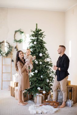 Üç kişilik mutlu aile, süslü Noel ağacı ve hediye kutularıyla rahat bir oturma odasında poz veriyor. Neşeli erkek ve kadın, neşeli atmosferde küçük kızlarıyla birlikte ebeveynliklerinin tadını çıkarıyorlar..