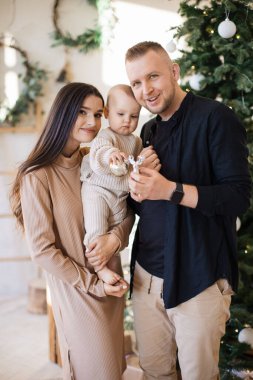 Güzel, küçük bir kızı olan, ışıklandırılmış ve Noel süslemeli bir odada poz veren sevimli ebeveynlerin ön görüntüsü. Gülümseyen beyaz aile yeni yıl havasında kameraya bakıyor.