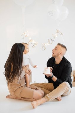 Kafkasyalı ebeveynler küçük kızlarını doğum günleriyle karşılamak için stüdyoda lezzetli pastalar, güzel hediyeler ve beyaz balonlar kullanıyorlar. Aile kavramı, yıldönümü ve kutlama.