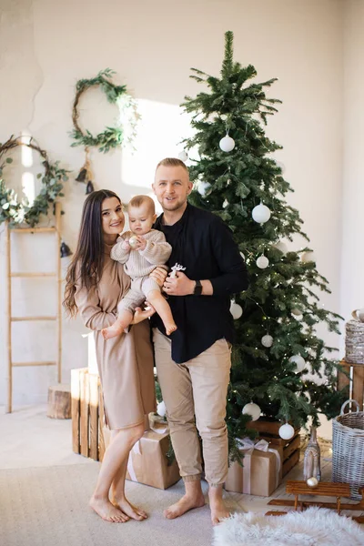 stock image Cheerful caucasian spouse holding beautiful little baby on hand while smiling and looking at camera. Beautiful Christmas tree with shiny garlands and balls placed behind at modern apartment.