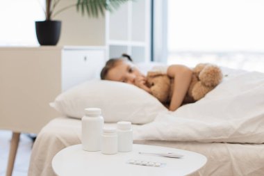 Beyaz komodinin yanında bir kavanoz hap ve küçük hasta kızın bulanık geçmişiyle yatak odasında duran termometre. Beyaz çocuk boğaz ağrısı çekerken oyuncak ayıyı kucaklıyor..