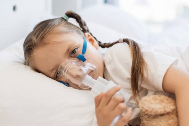 Küçük üzgün bir çocuğun portresi beyaz yatakta yatarken ve ayı oyuncağına sarılırken nebulizör maskesiyle ilaç soluyor. Kafkasyalı üzgün kız dinleniyor ve aydınlık yatak odasında tedavi oluyor..