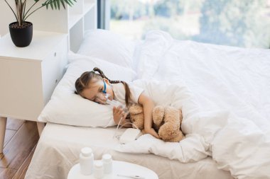 Grip olmuş güzel sarışın bir kız oyuncak ayıyla yatakta dinleniyor ve yüz maskesi takarak ilaç soluyor. Evde sağlık tedavisi kavramı.