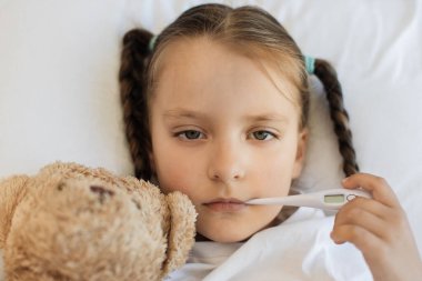 Hasta küçük kızın, yatakta ağzında dijital termometreyle sevgili oyuncak ayısına sarılışının en üst görüntüsü. Tıbbi hastalık ve ateş kavramı.